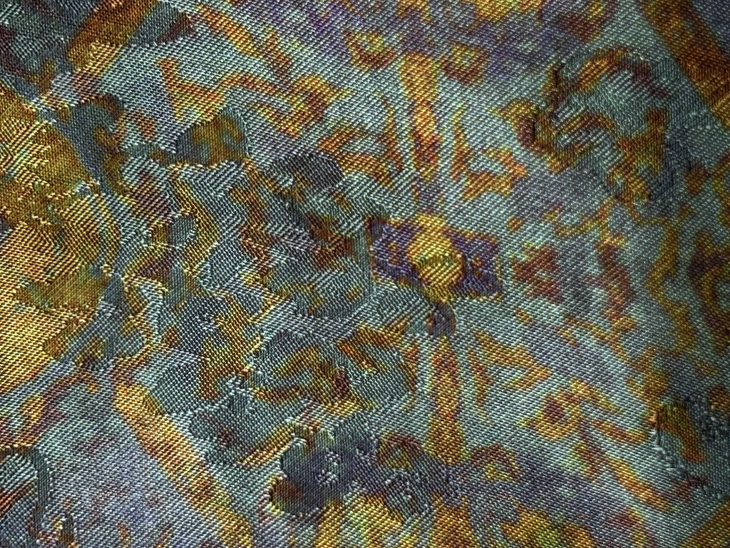 Close-up of a woven fabric with a dense, multicoloured abstract motif in teal, gray, gold, rust, and muted purple; the pattern appears irregular and layered across the surface.