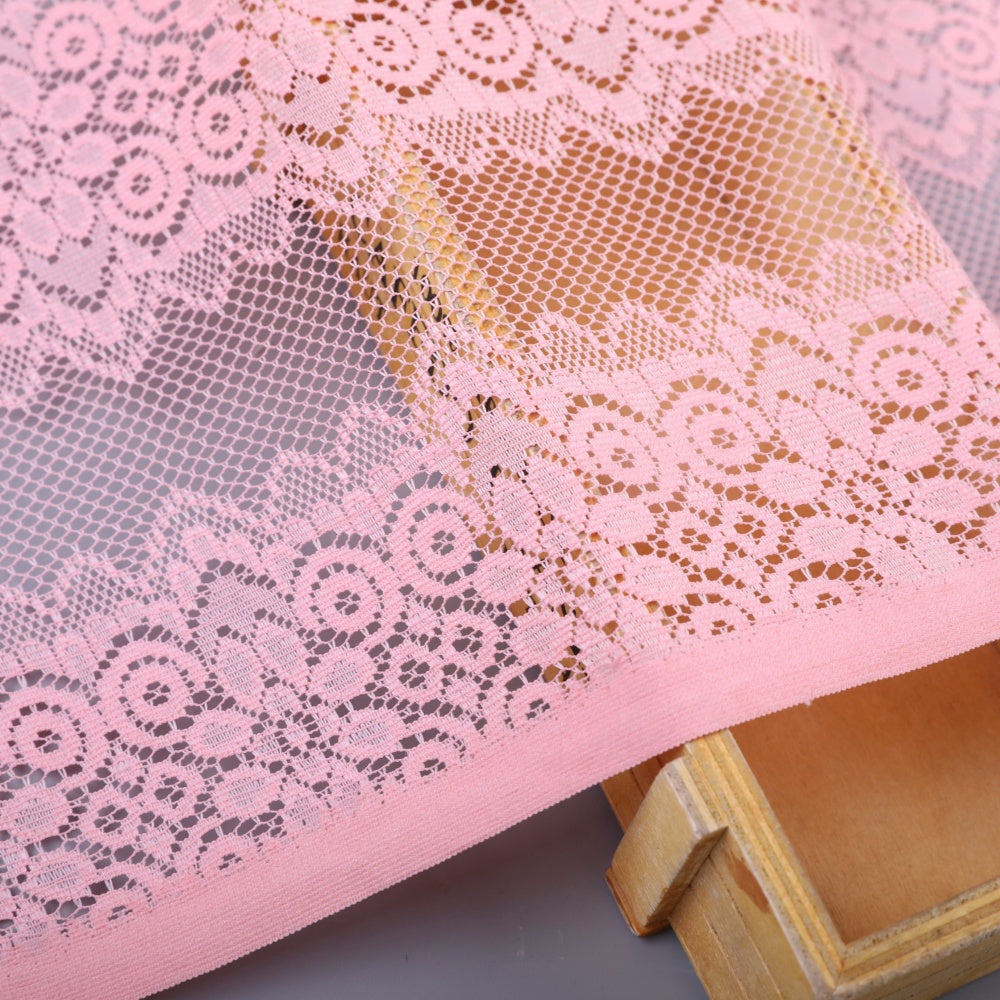 Close-up of pale pink lace fabric with open mesh and repeating floral and circular motifs, arranged over a wooden frame.