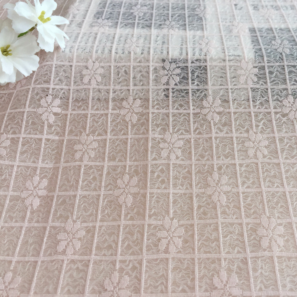 Close-up of pale blush lace with a repeating grid of embroidered flowers and wavy lines; white flowers appear at the upper left.