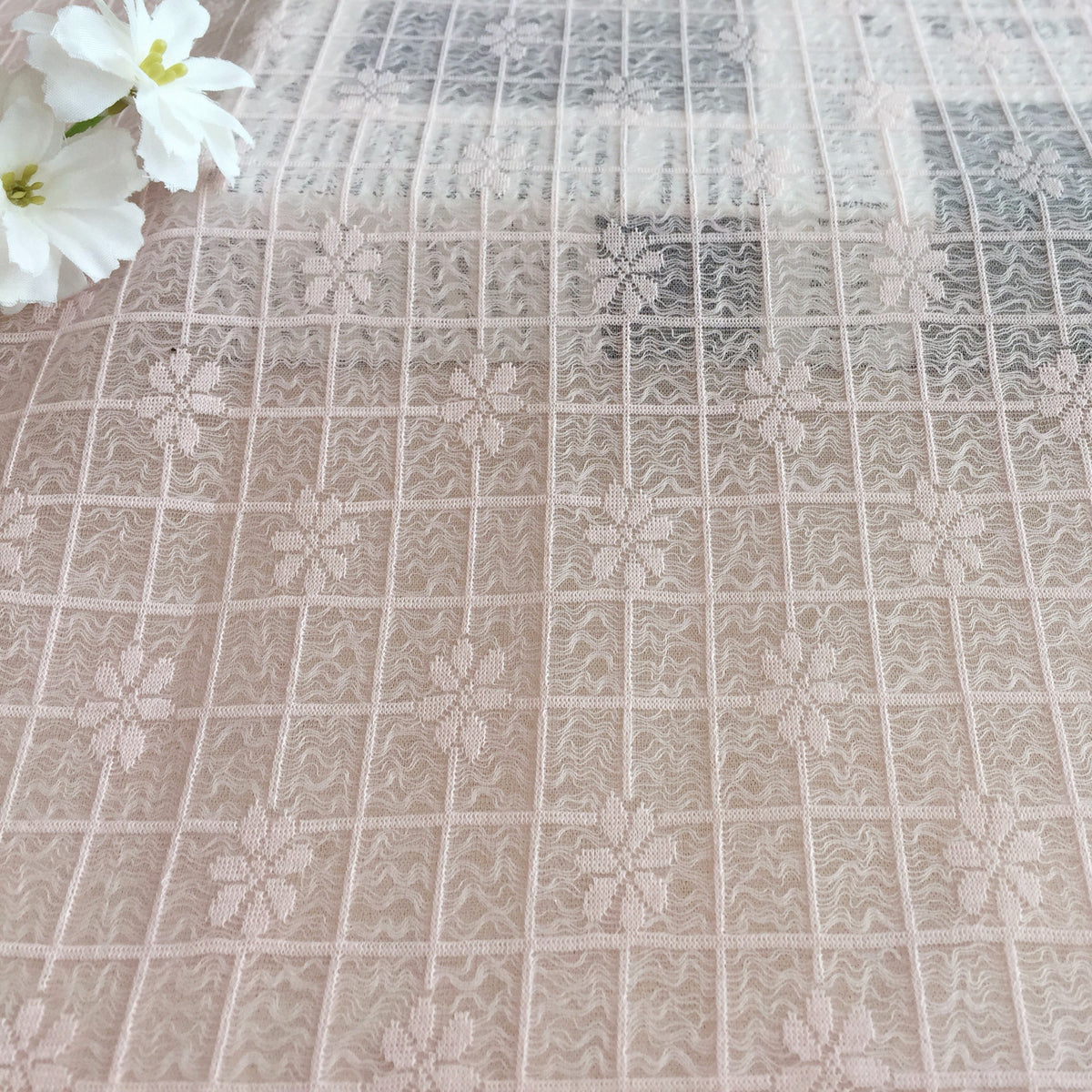 Close-up of pale blush lace with a repeating grid of embroidered flowers and wavy lines; white flowers appear at the upper left.