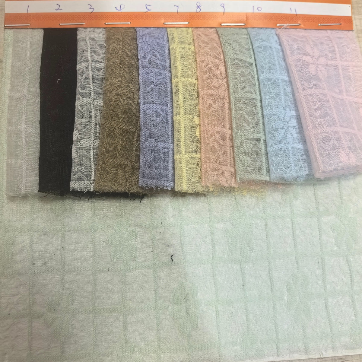 Close-up of overlapping rectangular fabric swatches in muted gray, black, brown, lavender, yellow, peach, sage, blue, and pink, with repeating wavy lace-like motifs and panel borders; a pale patterned cloth lies underneath.