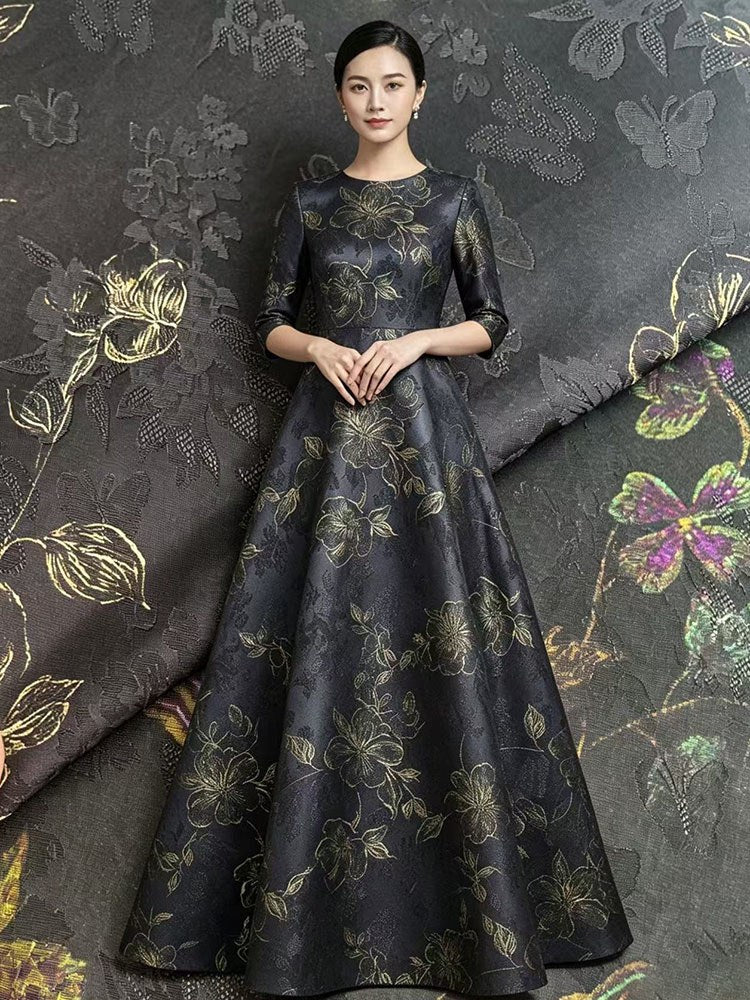 A full-length view of a woman wearing a dark charcoal floor-length dress with gold and muted green floral motifs; matching patterned fabric is displayed behind her.