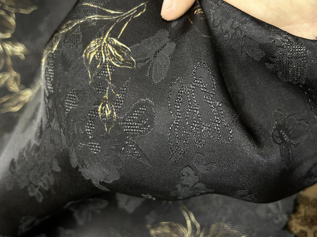Close-up of black cloth with a subtle tonal floral and leaf pattern, plus a few gold-toned embroidered stems; the fabric is folded and held open by a hand.