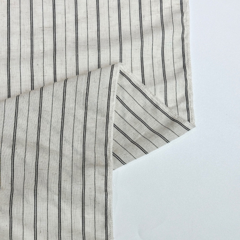 An off-white fabric with narrow black vertical stripes is laid flat, with one corner folded back to show the stripes running diagonally on the fold and a stitched edge.