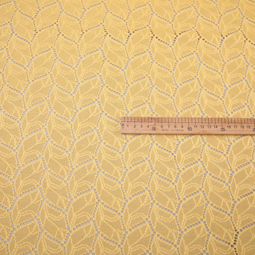 Close-up of golden yellow lace fabric with repeating openwork leaf motifs arranged in diagonal rows; a wooden centimeter ruler lies across the fabric.
