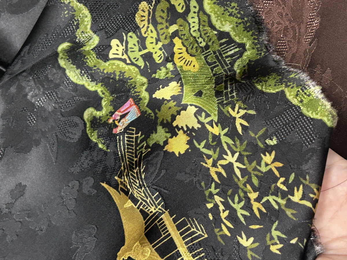 Close-up of black patterned cloth with olive green, yellow, and gold floral and leafy motifs, plus a small red accent; the fabric is draped and folded.