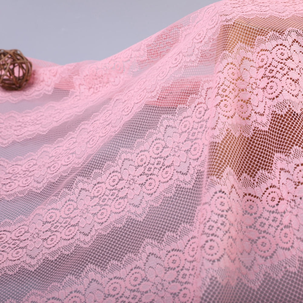 Close-up of pale pink lace fabric draped in curved folds, with repeating floral motifs and open mesh sections; a small brown woven ball sits near the upper left.