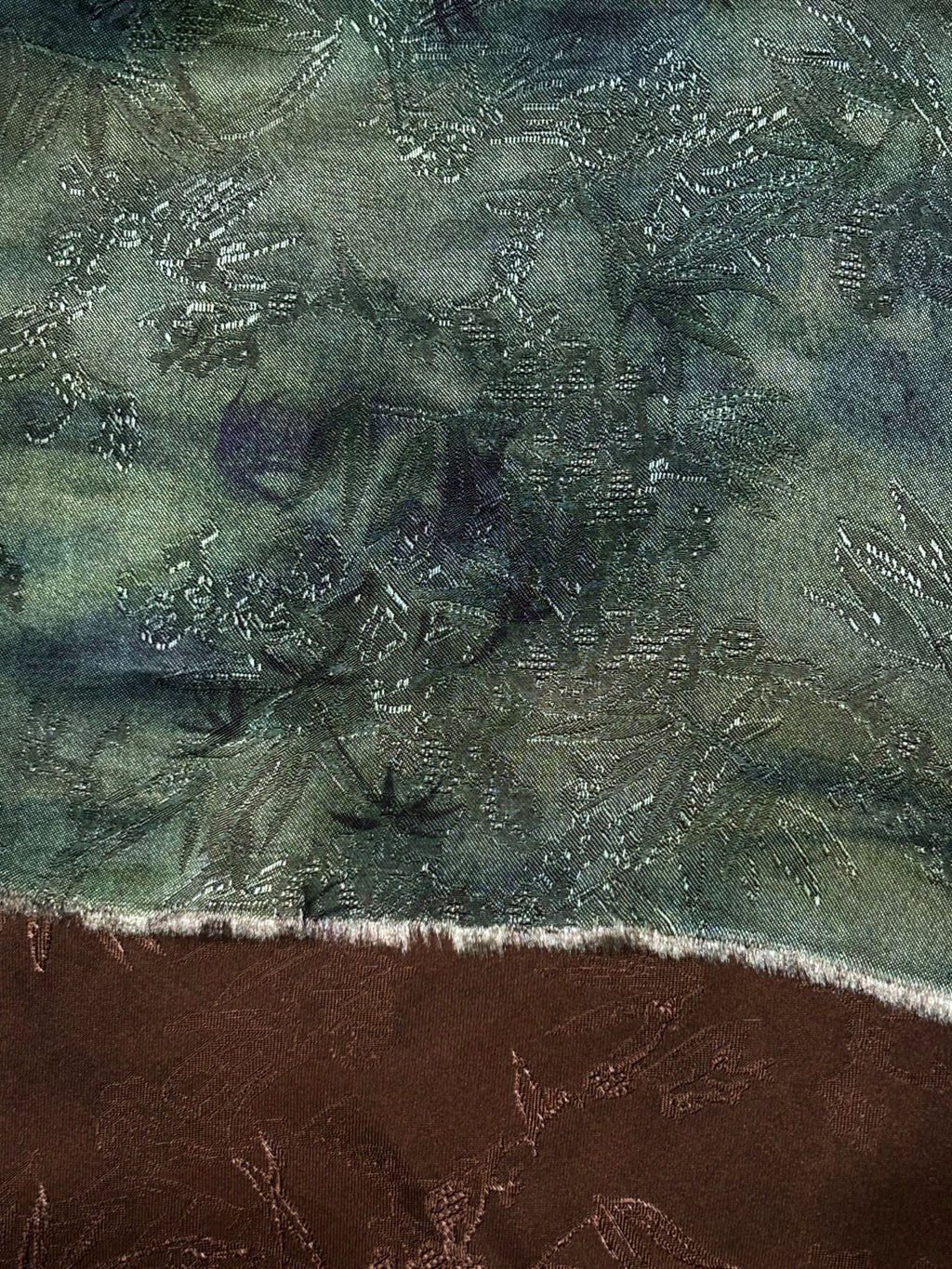 Close-up of a dark green woven fabric with an intricate, irregular pattern of pale stitched lines and darker motifs; a curved edge with short white fringe separates it from brown fabric below.
