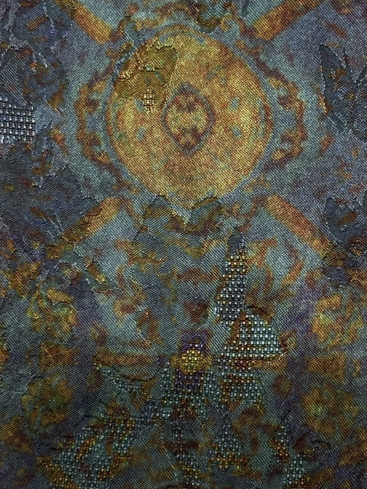 Close-up of dark blue-gray fabric with a large, ornate gold and muted brown circular motif, surrounded by smaller geometric and floral details; the design is interrupted by scattered rows of reflective square accents.