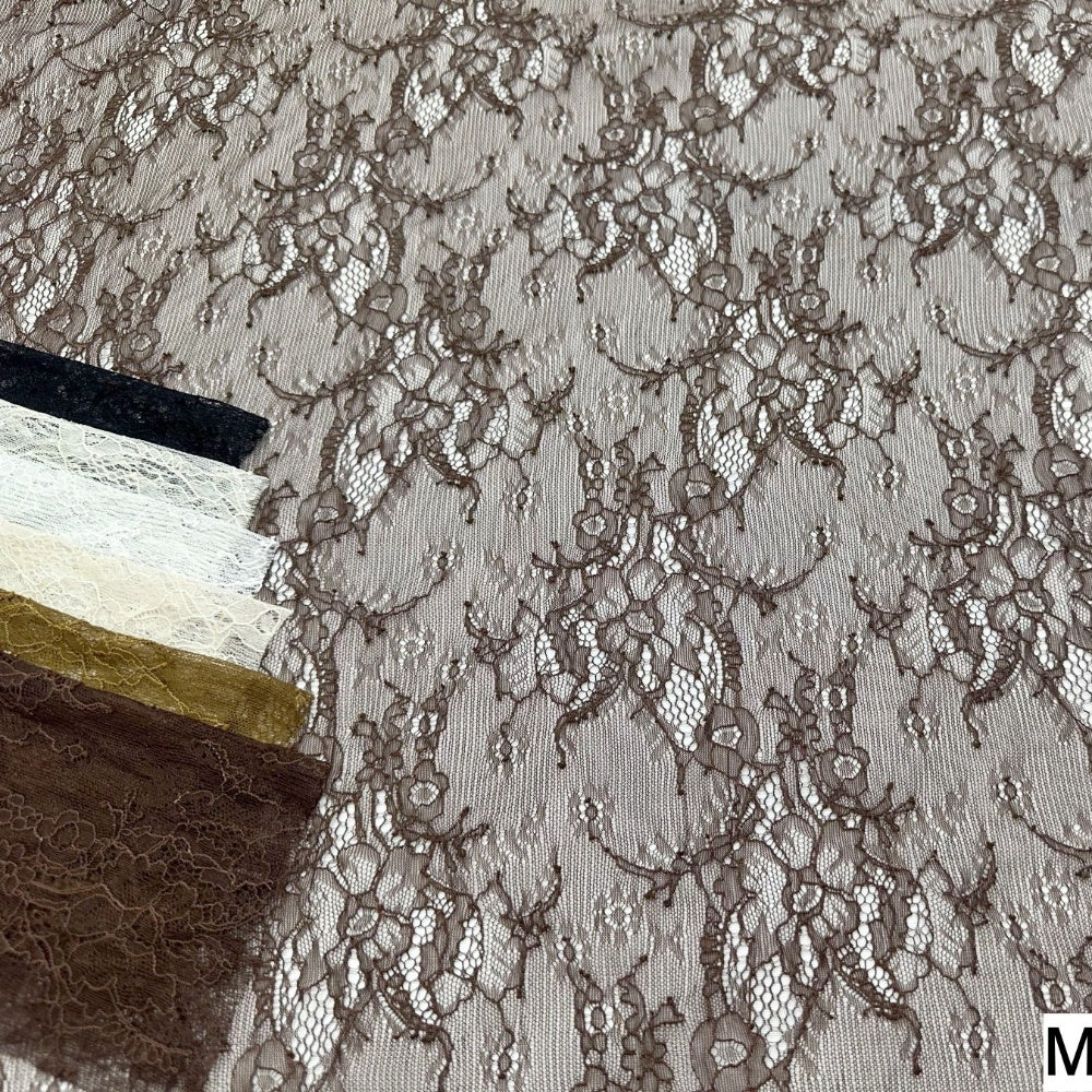 Brown floral lace fabric is laid flat, with repeating openwork flowers and curling vines across a light taupe ground; dark navy, ivory, gold, and brown fabric swatches overlap along the left edge.
