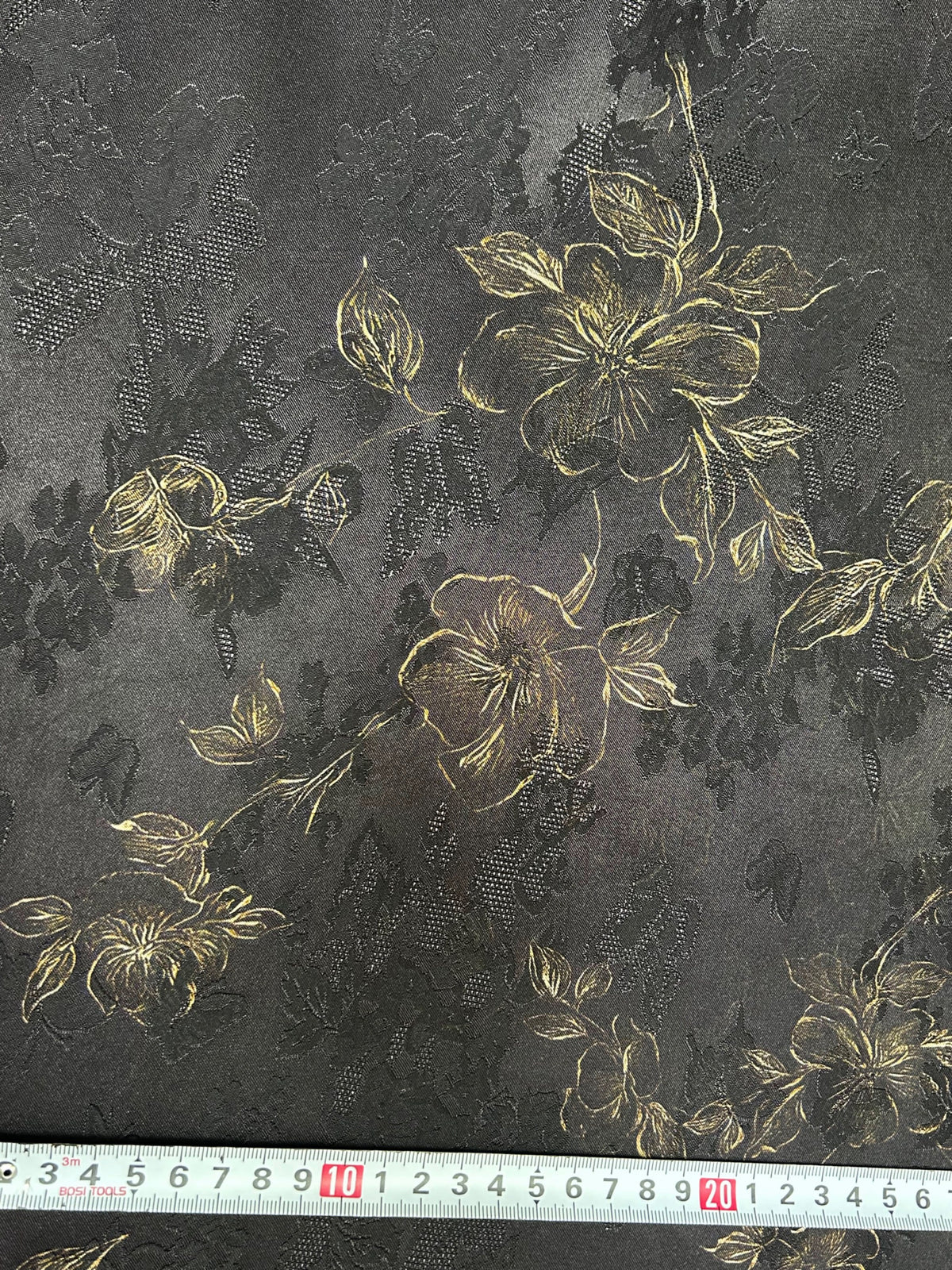 Close-up of a charcoal gray fabric swatch with scattered black floral silhouettes and fine gold line-drawn flowers, leaves, and buds; a measuring tape runs along the bottom edge.