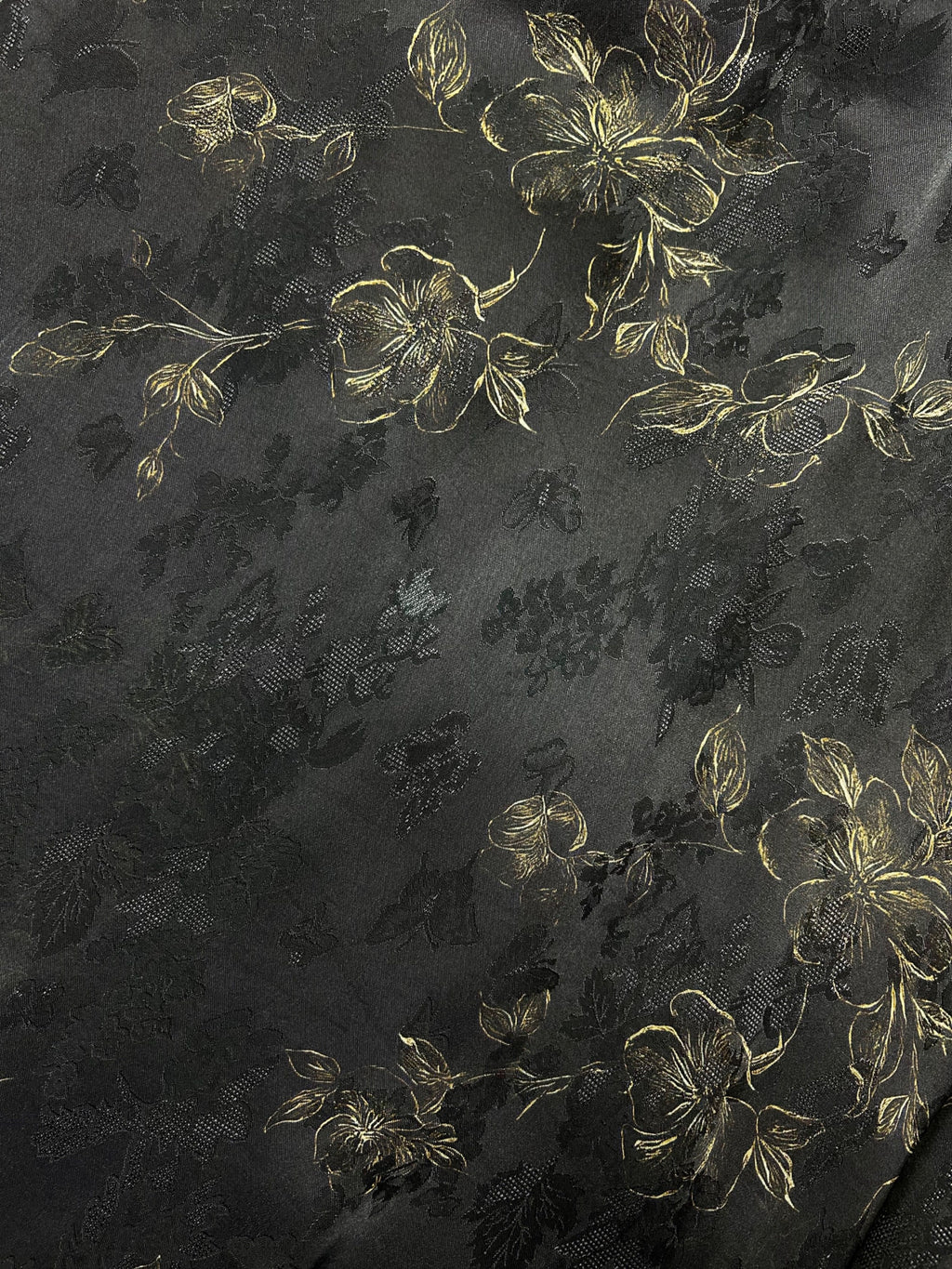 Close-up of dark charcoal fabric with a subtle black floral pattern and metallic gold flower outlines and leaves arranged in diagonal clusters.