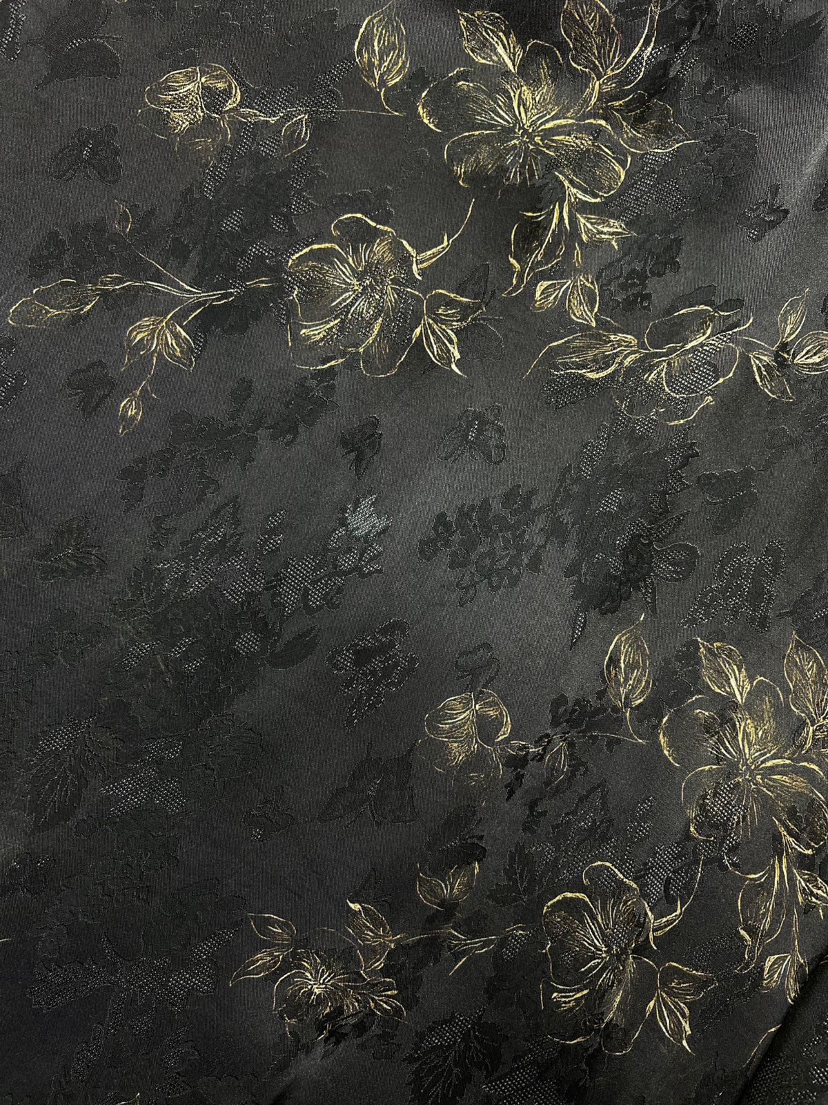 Close-up of dark charcoal fabric with a subtle black floral pattern and metallic gold flower outlines and leaves arranged in diagonal clusters.
