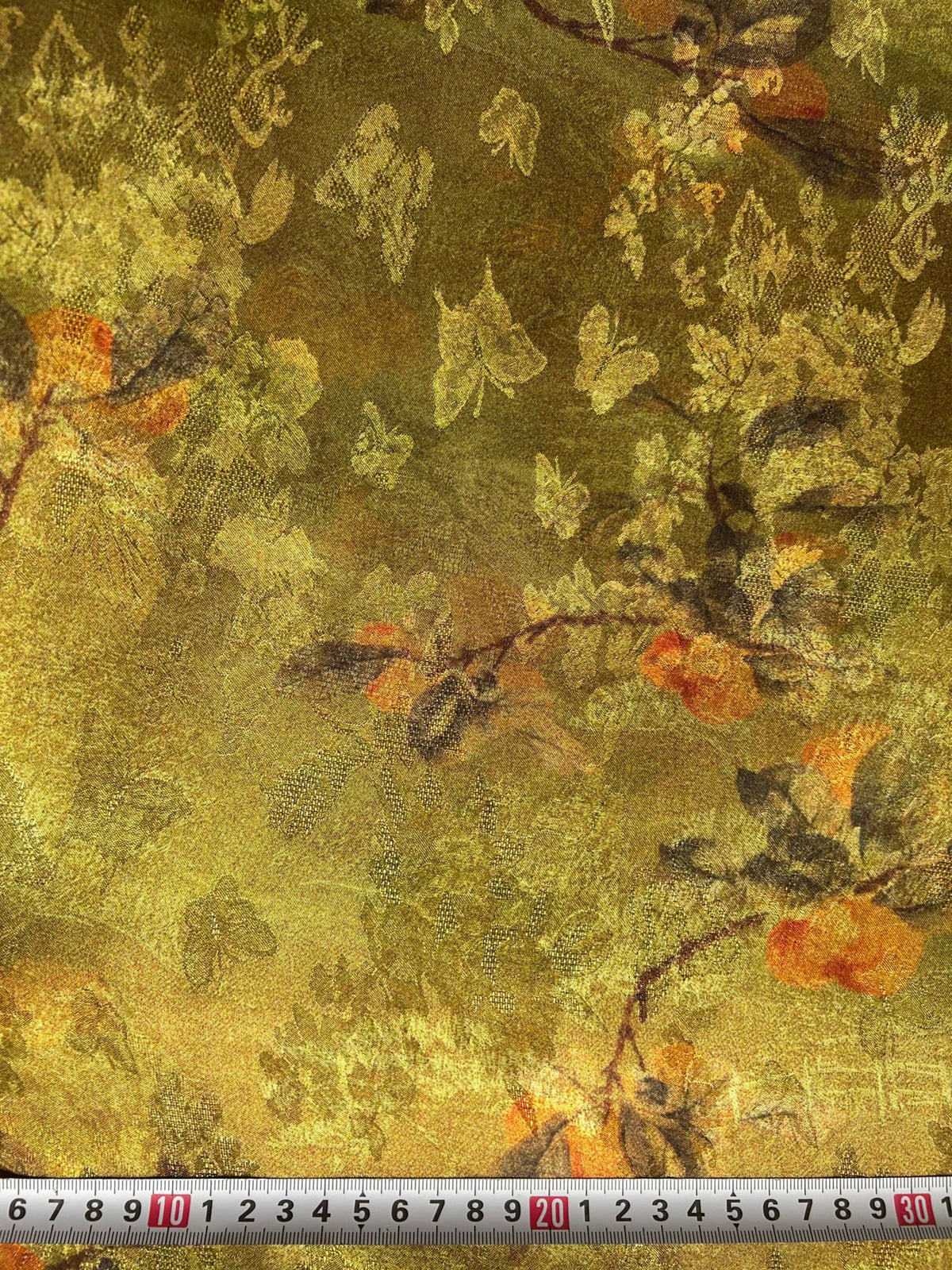 Close-up of a golden olive fabric swatch with a dense, irregular floral and leafy motif in pale yellow, orange, brown, and dark green; a measuring tape runs along the bottom edge.
