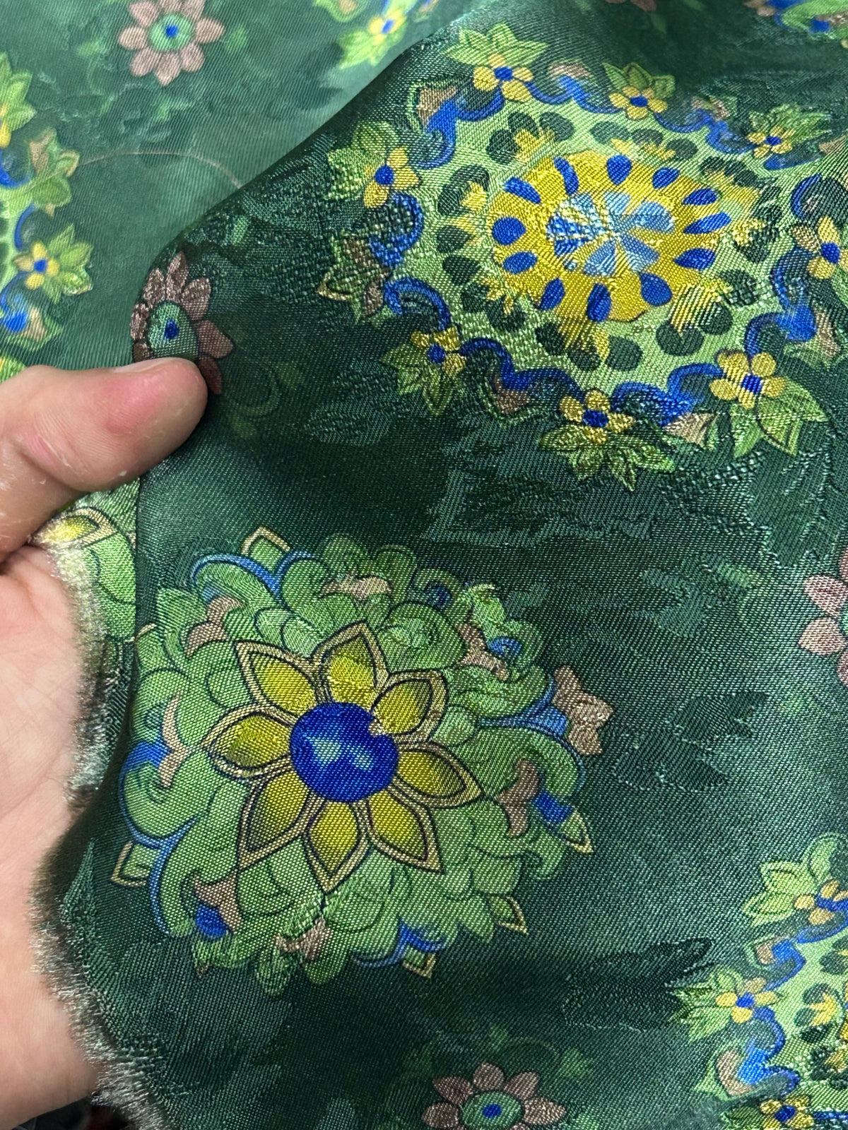 Close-up of a folded dark green cloth with dense floral motifs in yellow, blue, light green, and muted pink; a hand holds one edge.