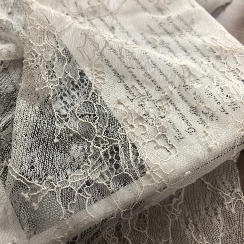 Ivory lace fabric is loosely draped in overlapping folds, showing an open net ground with raised floral and leafy motifs and small scattered knots.