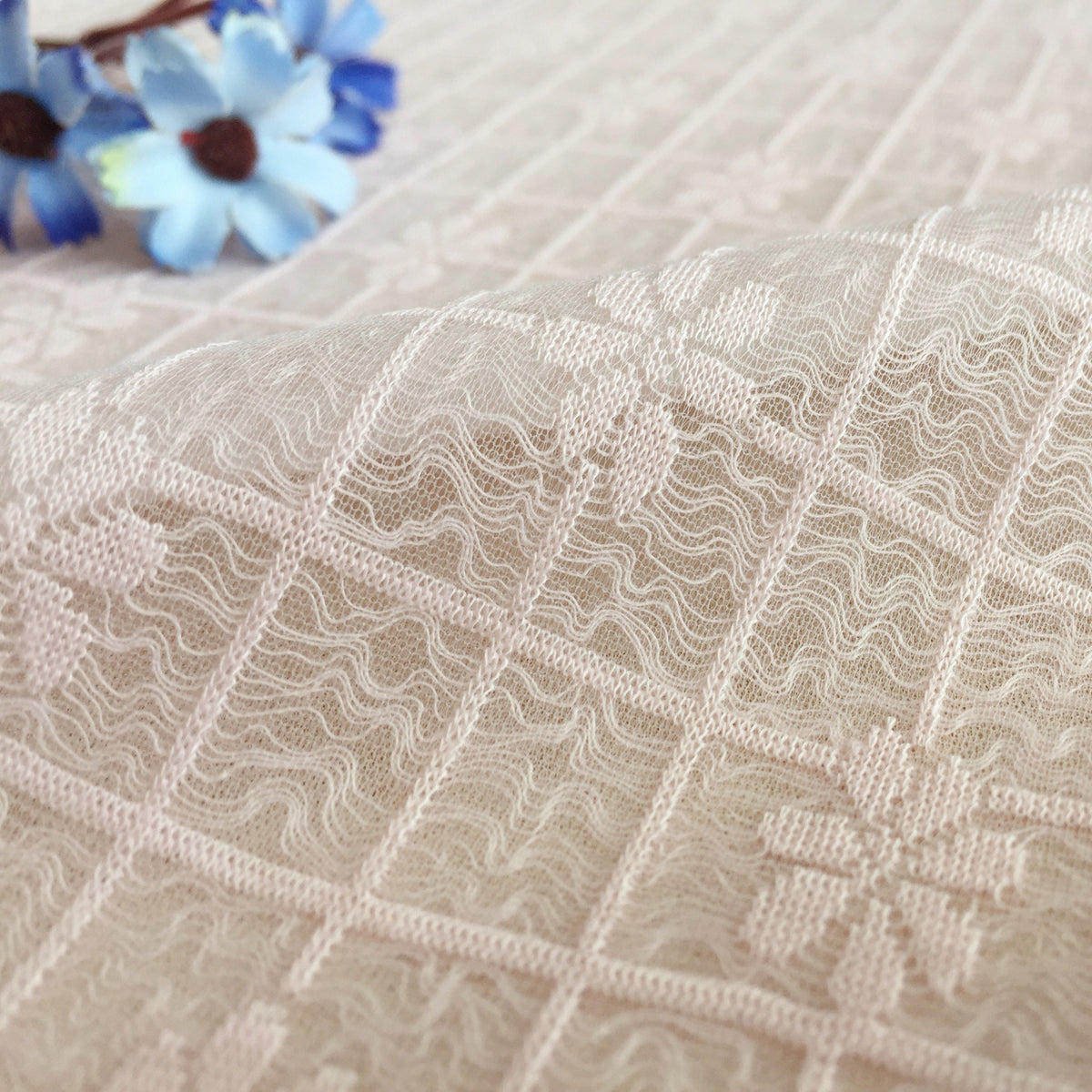 Close-up of pale ivory lace with repeating rectangular panels filled with wavy lines, draped across the foreground; blurred blue flowers appear in the upper left.