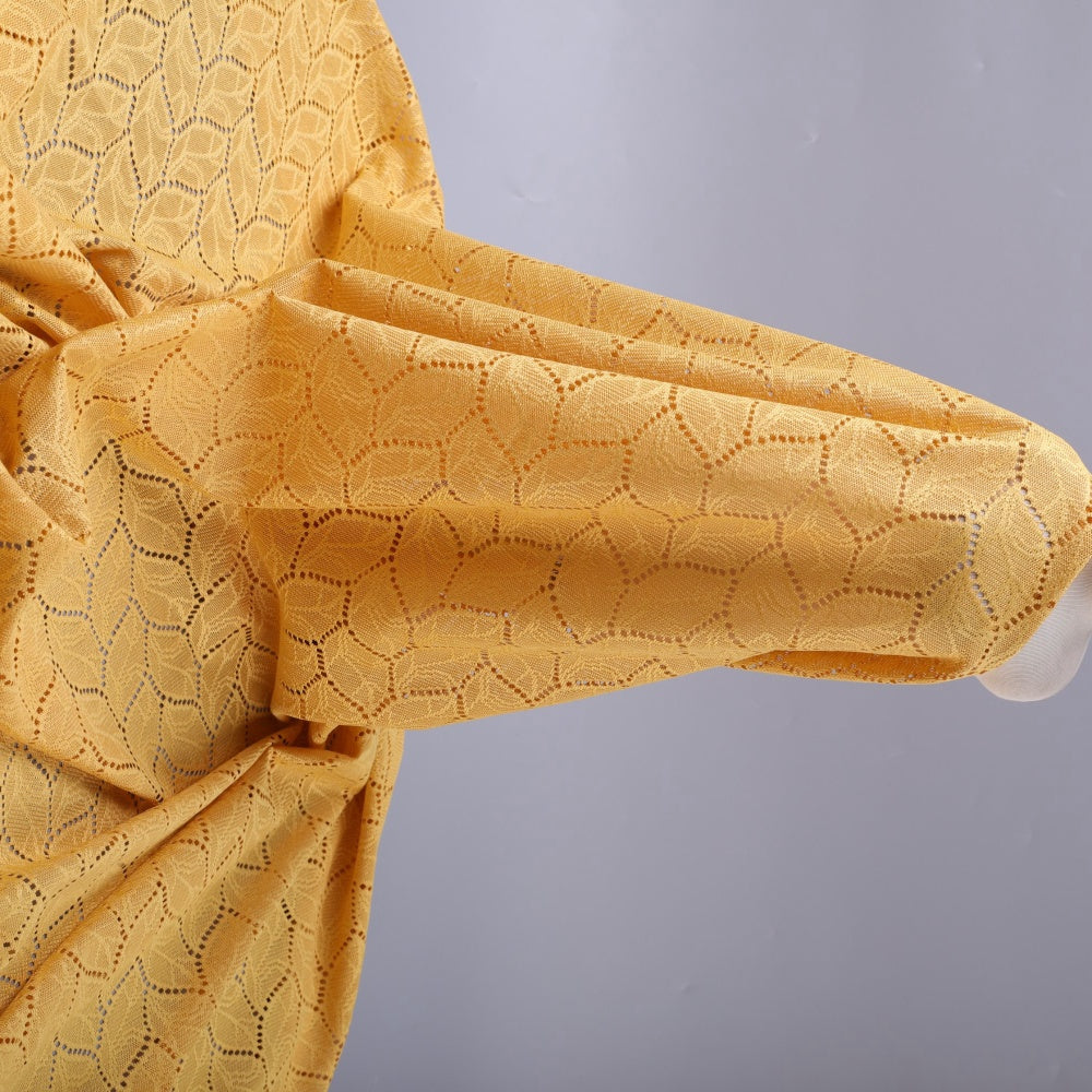 Close-up of mustard yellow cloth with an openwork repeating leaf motif, draped in overlapping folds across a pale gray background.