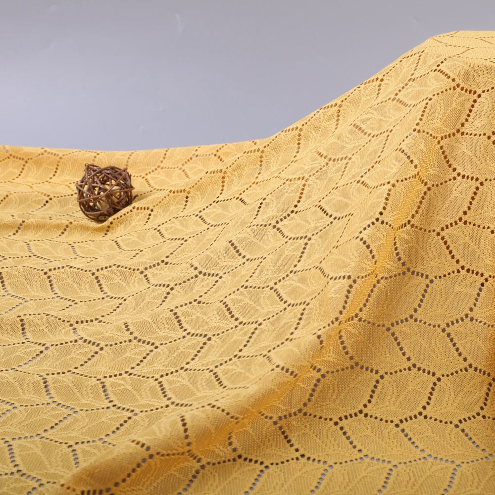 Close-up of golden yellow cloth draped in curved folds, with a repeating openwork pattern of wavy dotted lines and leaf shapes; a small brown woven ball rests near the upper left.