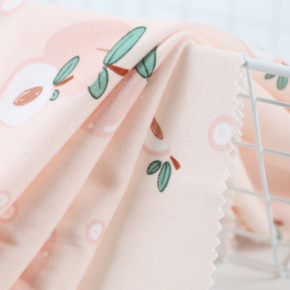 Close-up of pale peach cloth draped in curved folds, with scattered white and rust-colored shapes and small green leaf motifs; a zigzag-cut edge hangs beside a white wire rack.