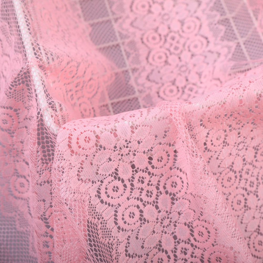Close-up of pale pink lace fabric with repeating floral and geometric motifs, draped in overlapping folds.