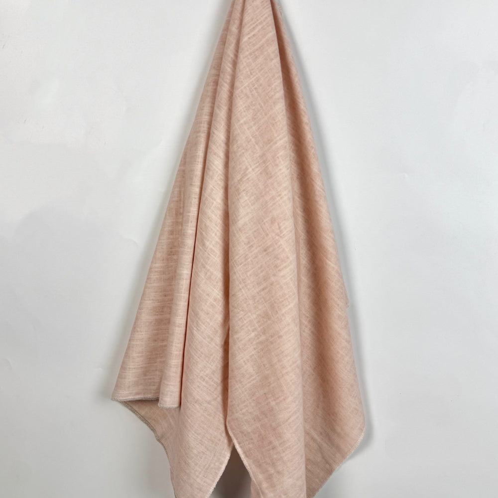Pale blush pink fabric hangs in long, flowing folds against a white background, showing a subtle crosshatched texture and narrow finished edges.