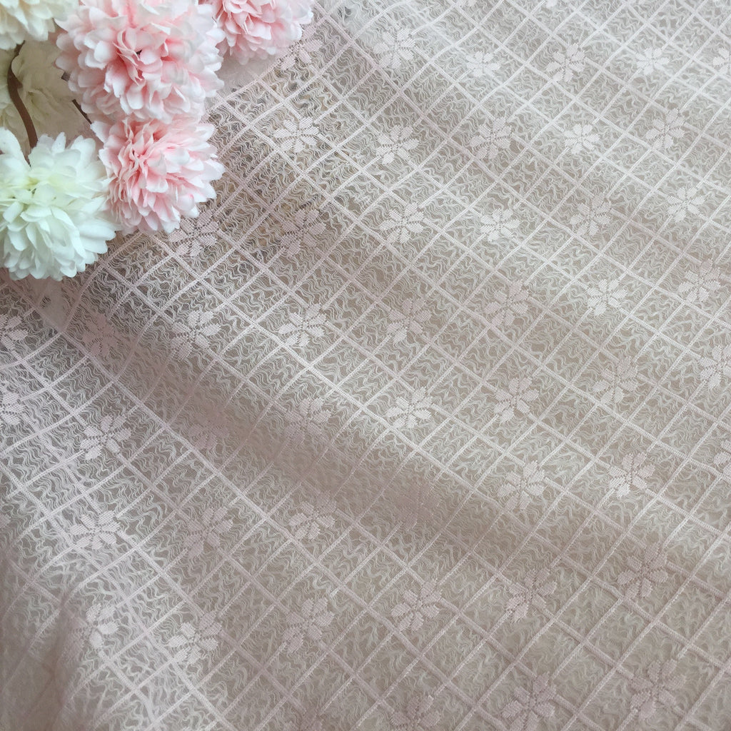 Close-up of pale beige lace with a diagonal diamond grid and floral motifs; pale pink and white flowers cluster along the upper left edge.