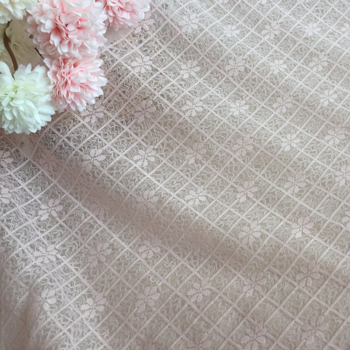 Close-up of pale beige lace with a diagonal diamond grid and floral motifs; pale pink and white flowers cluster along the upper left edge.